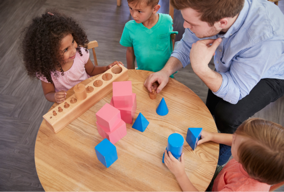 Children working with Montessori wooden blocks and geometric shapes alongside a teacher at a classroom table.