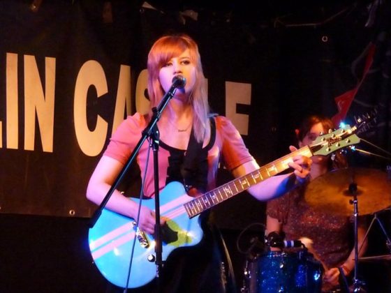 Marisa Levenson performing with a blue electric guitar and singing into a microphone with The Common Earth