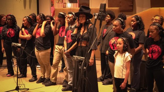 Snooknuk standing with a group of young musicians wearing headphones in a music studio